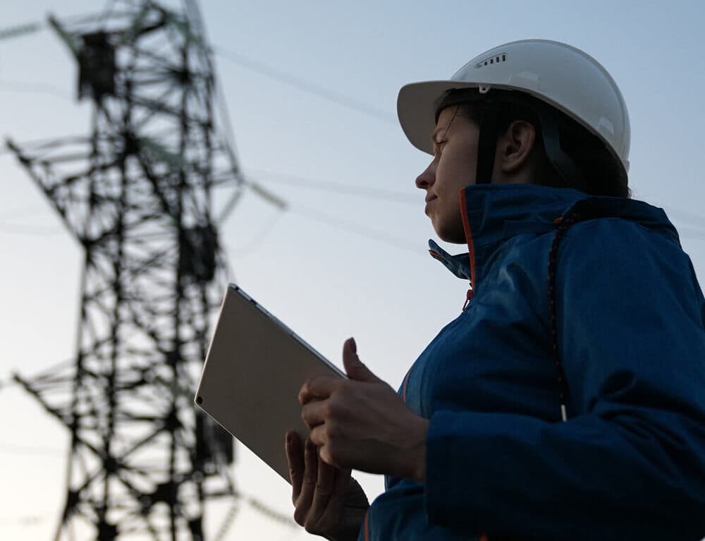 Kvinna klädd i arbetskläder och skyddshjälm, som håller en surfplatta och granskar en kraftledningsstolpe i bakgrunden. Scenen utspelar sig vid skymning, vilket belyser arbetet inom energisektorn och infrastrukturen för eldistribution.